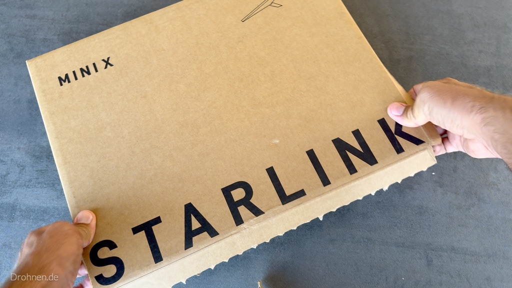 Starlink-Mini-X-Karton auf einem Tisch mit geöffneter Aufreißkante und Händen am Deckel.