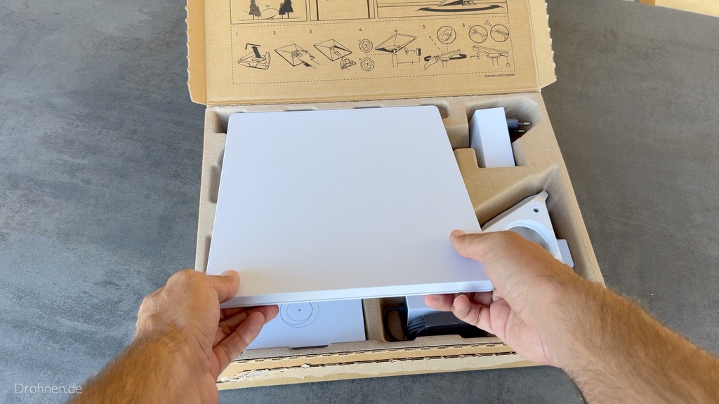 Zwei Hände heben das weiße Starlink-Mini-Terminal aus dem geöffneten Karton.