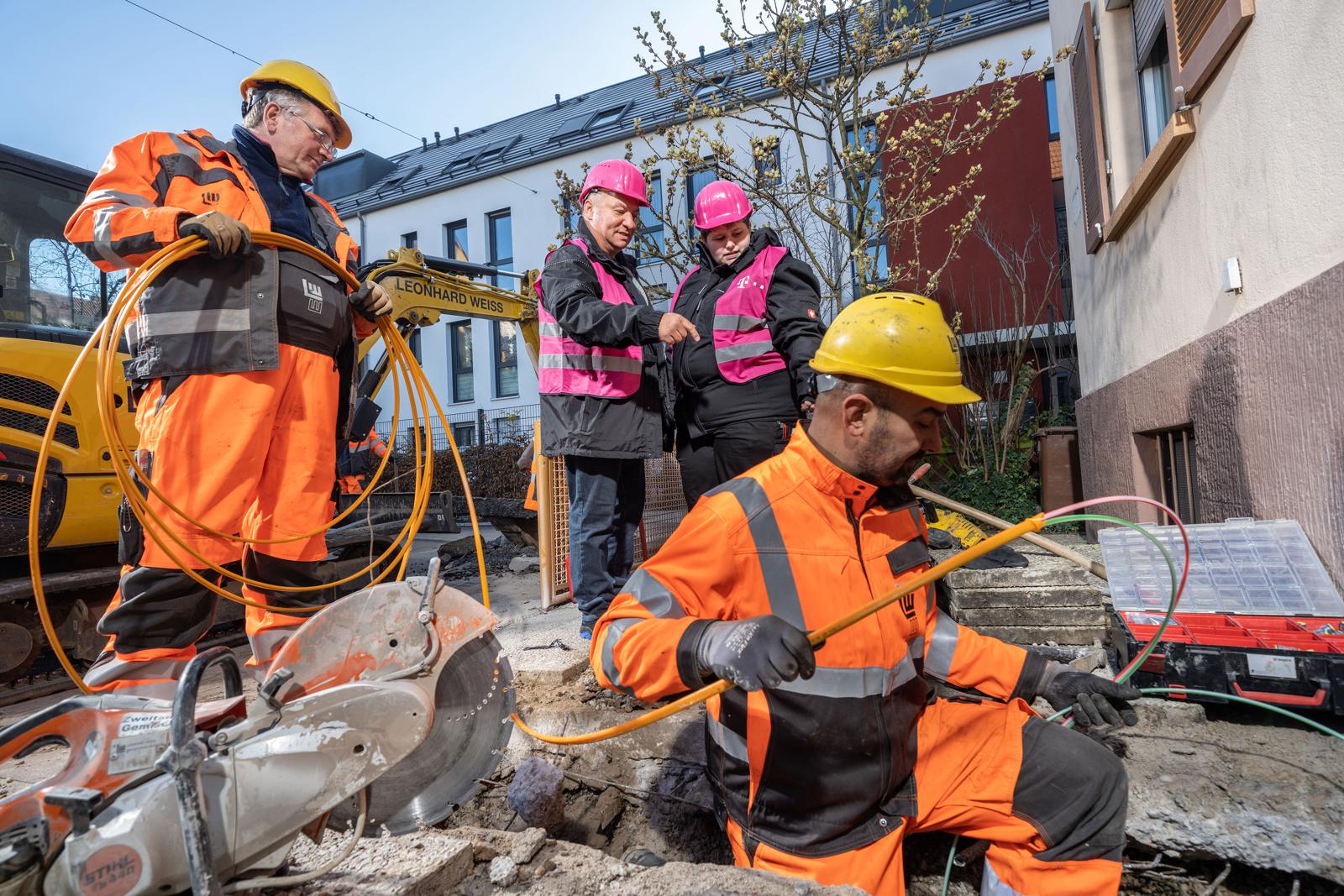 Bauarbeiter in orangefarbener Warnkleidung verlegen auf einer Straßenbaustelle Glasfaserkabel, dahinter zwei Baubegleiterinnen in pinkfarbenen Westen