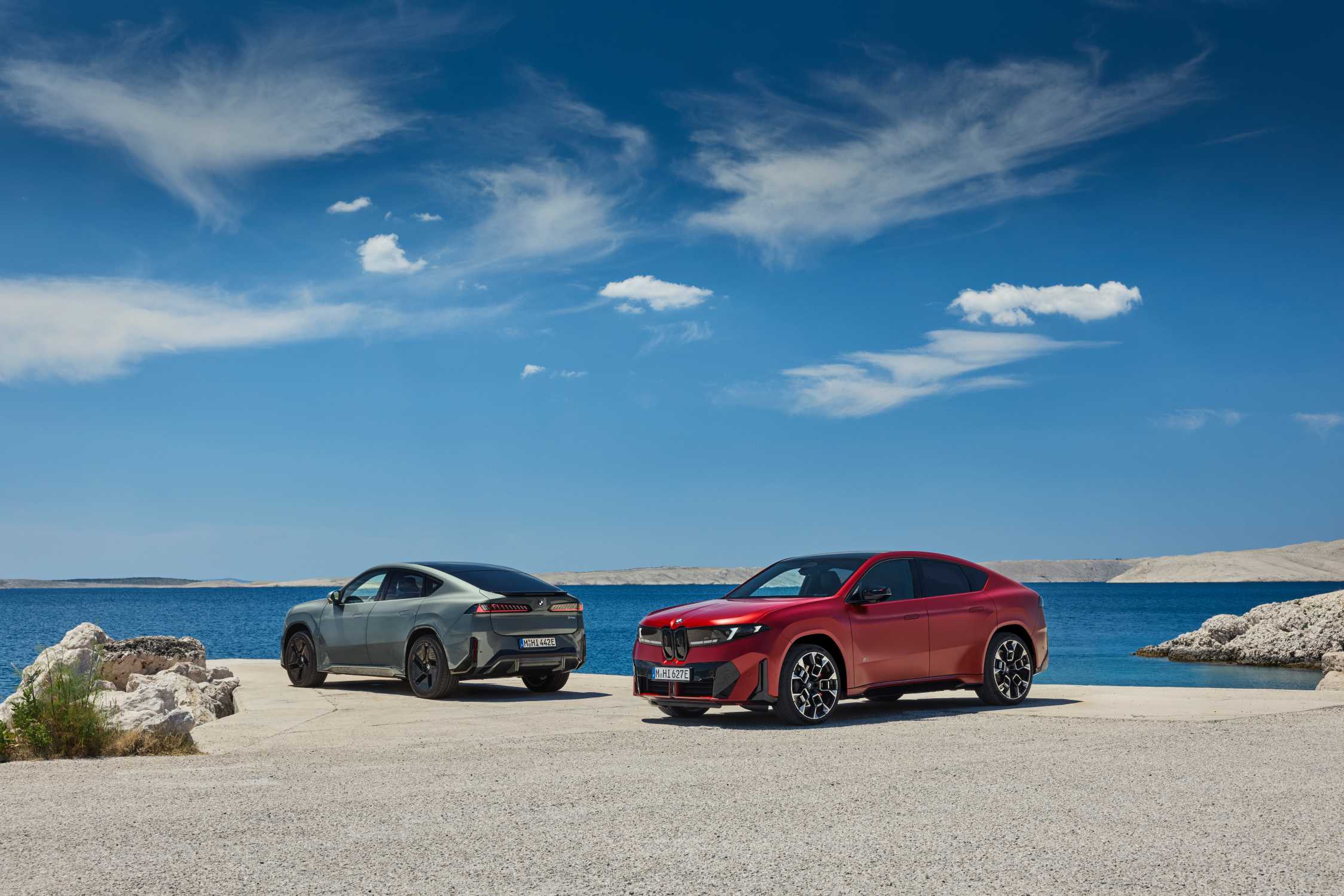 Originales BMW-Pressebild mit zwei BMW iX4 an einer Küstenstraße, einem roten Fahrzeug im Vordergrund und einem grauen dahinter.
