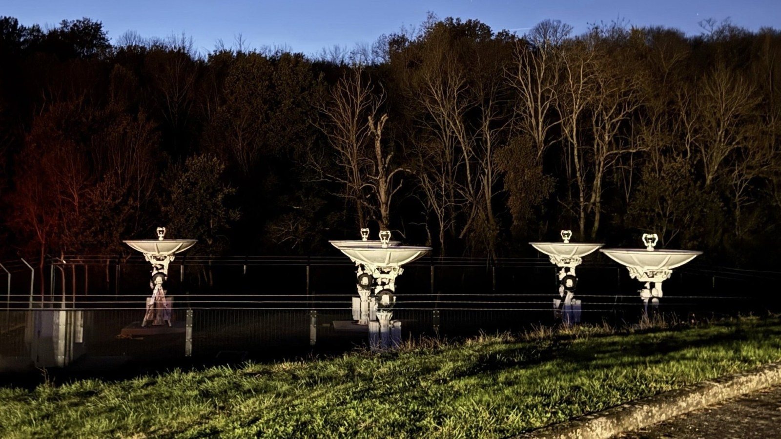 Amazon-Leo-Bodenstation mit fünf beleuchteten Parabolantennen hinter einem Zaun vor einem Waldrand in der Abenddämmerung