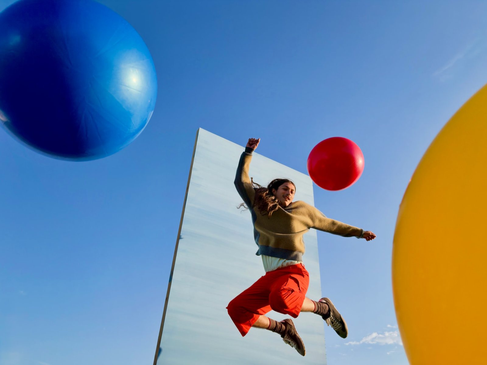 Beispielfoto der Hauptkamera: Eine Person springt vor blauem Himmel zwischen großen bunten Kugeln und einer Spiegelwand