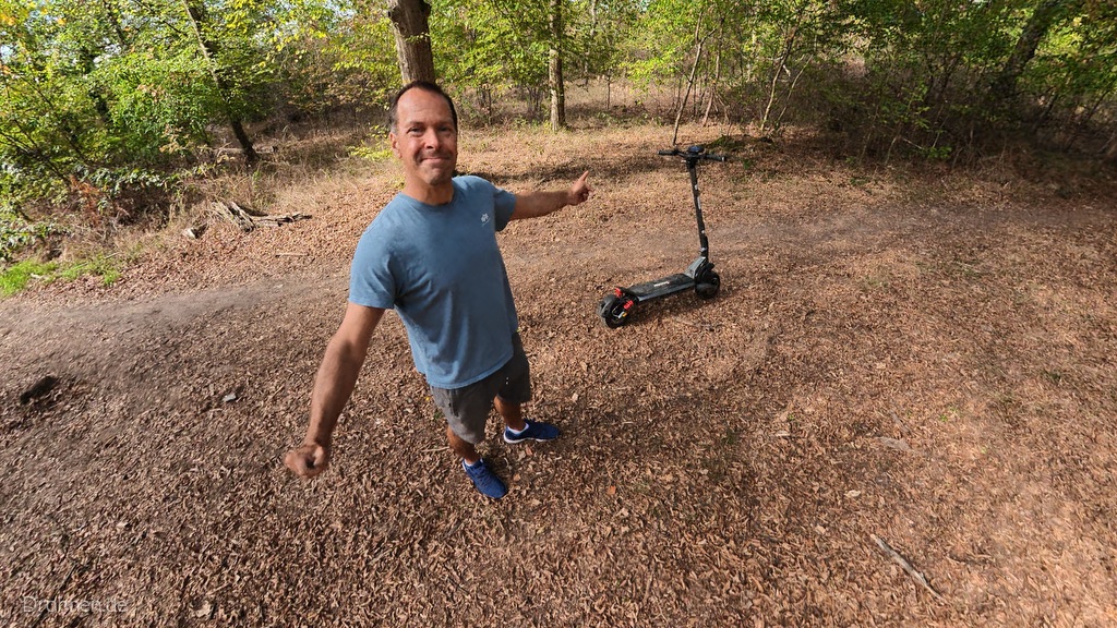 Tester zeigt auf den iScooter iX7 Pro, der auf einer Waldlichtung abgestellt ist